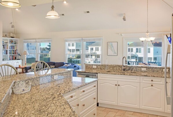 A modern kitchen with granite countertops, white cabinetry, and a large island. There are multiple windows providing natural light and views of the neighborhood. - Dog Daze: Private Pool & Spa, Dog-friendly, Club Access, Ocean & Sound Views (Corolla)