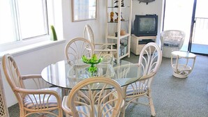 A dining area with a glass table surrounded by wicker chairs, a television, and a bookshelf. - Even Keel 404-Oceanfront 75th St, Elev, W/D, AC (Ocean City)