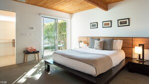 Un dormitorio moderno con techo de madera, una cama grande con muchas almohadas, un banco con toallas y un ventanal corredero que da a un espacio exterior.