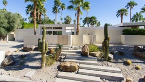 Un edificio moderno con diseño minimalista, rodeado de un jardín desértico con cactus y palmeras.