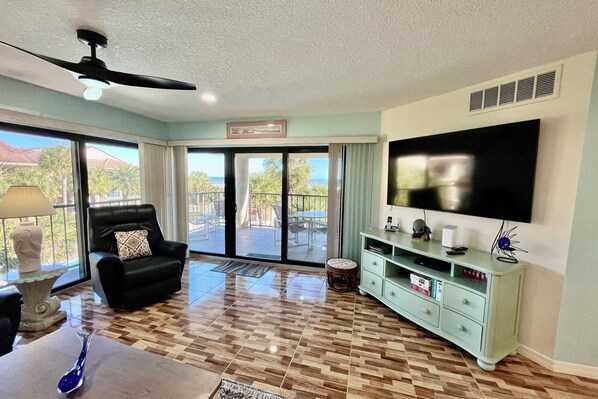 A spacious living room with a large TV, a ceiling fan, a black leather chair, and a view through the glass doors to the outside.