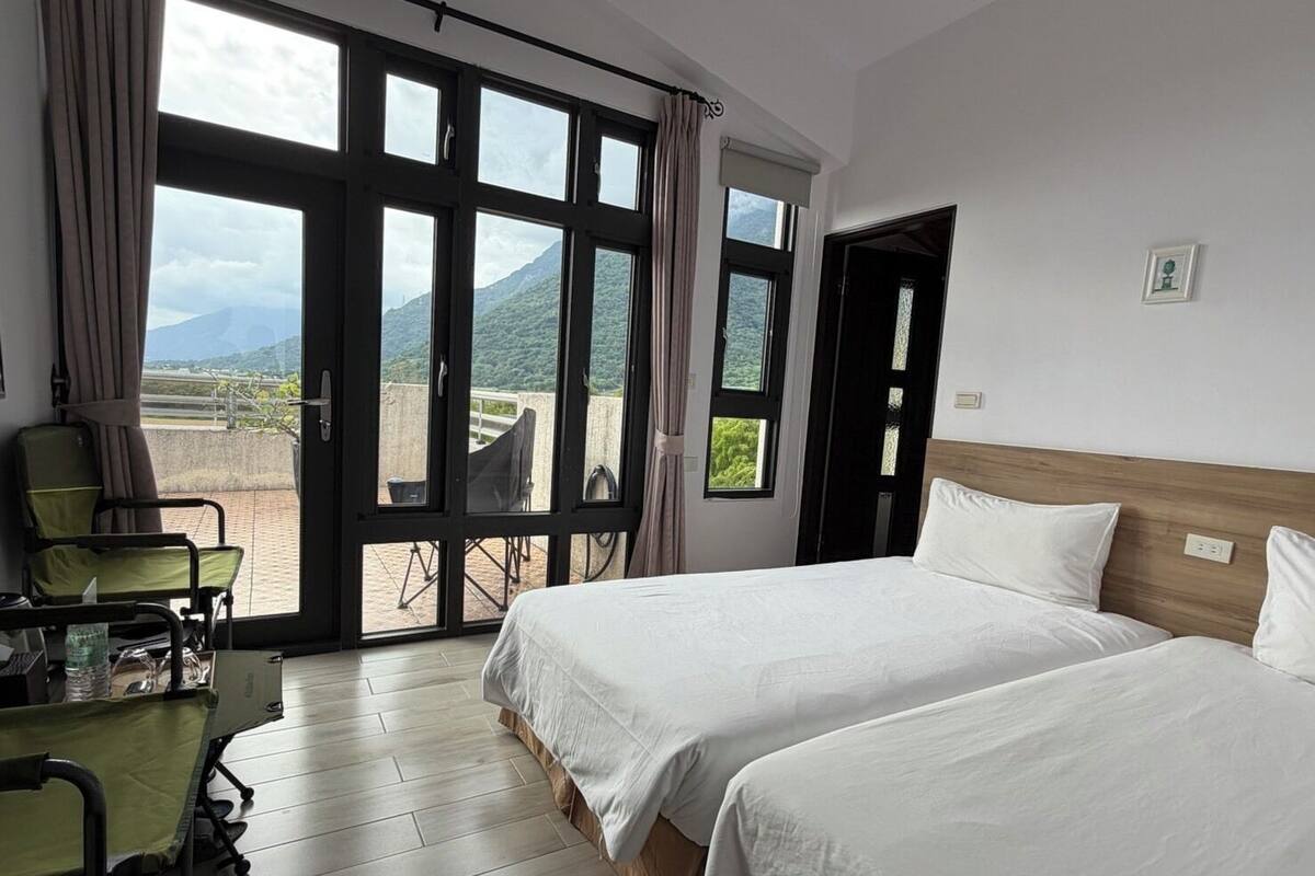 Habitación de hotel con dos camas, una silla de ruedas y vistas a montañas a través de amplias ventanas.