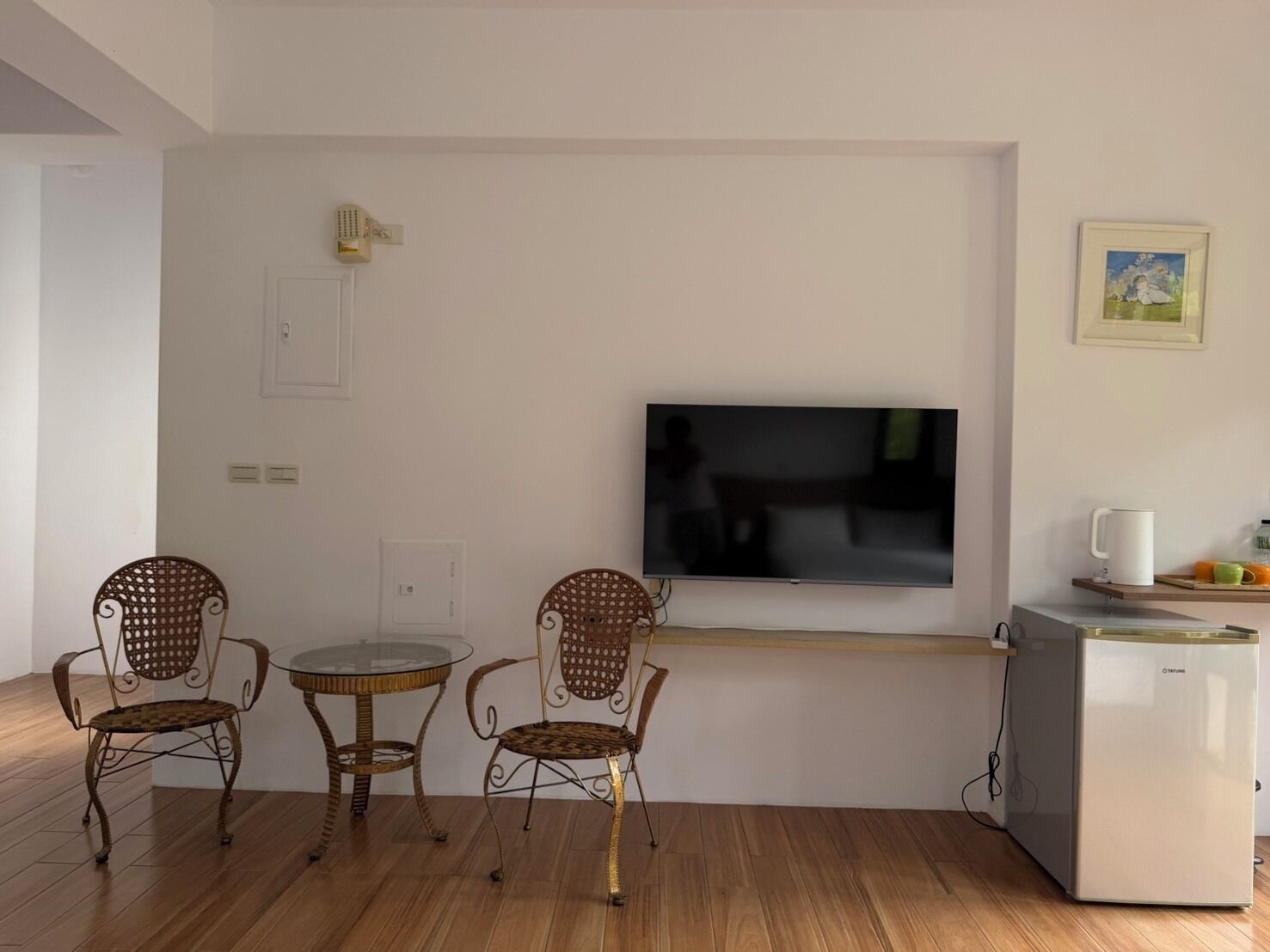 A small, modern living room with a flat-screen TV mounted on the wall, two wicker chairs, a small round table, and a white refrigerator.