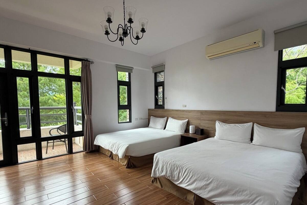 Habitación de hotel con dos camas, un candelabro, equipo de aire acondicionado y grandes ventanas con vistas a zonas verdes.