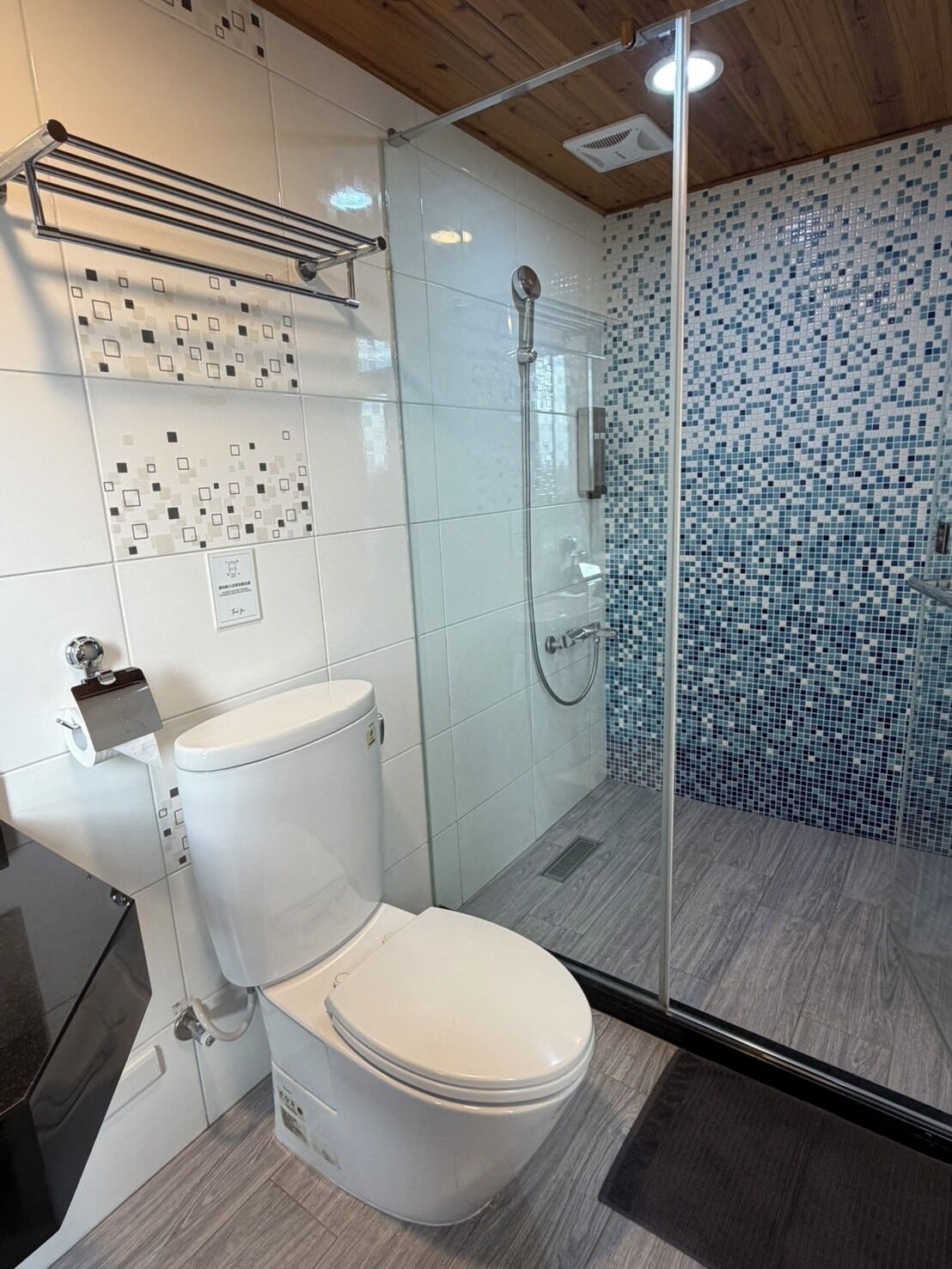 Un baño moderno con inodoro, ducha con mampara de vidrio y pared de azulejos con diseño tipo mosaico.
