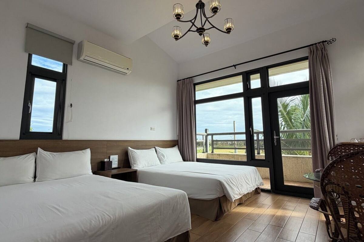 Habitación de hotel con dos camas, una araña, unidad de aire acondicionado, ventana con cortinas y un balcón con una silla de mimbre.