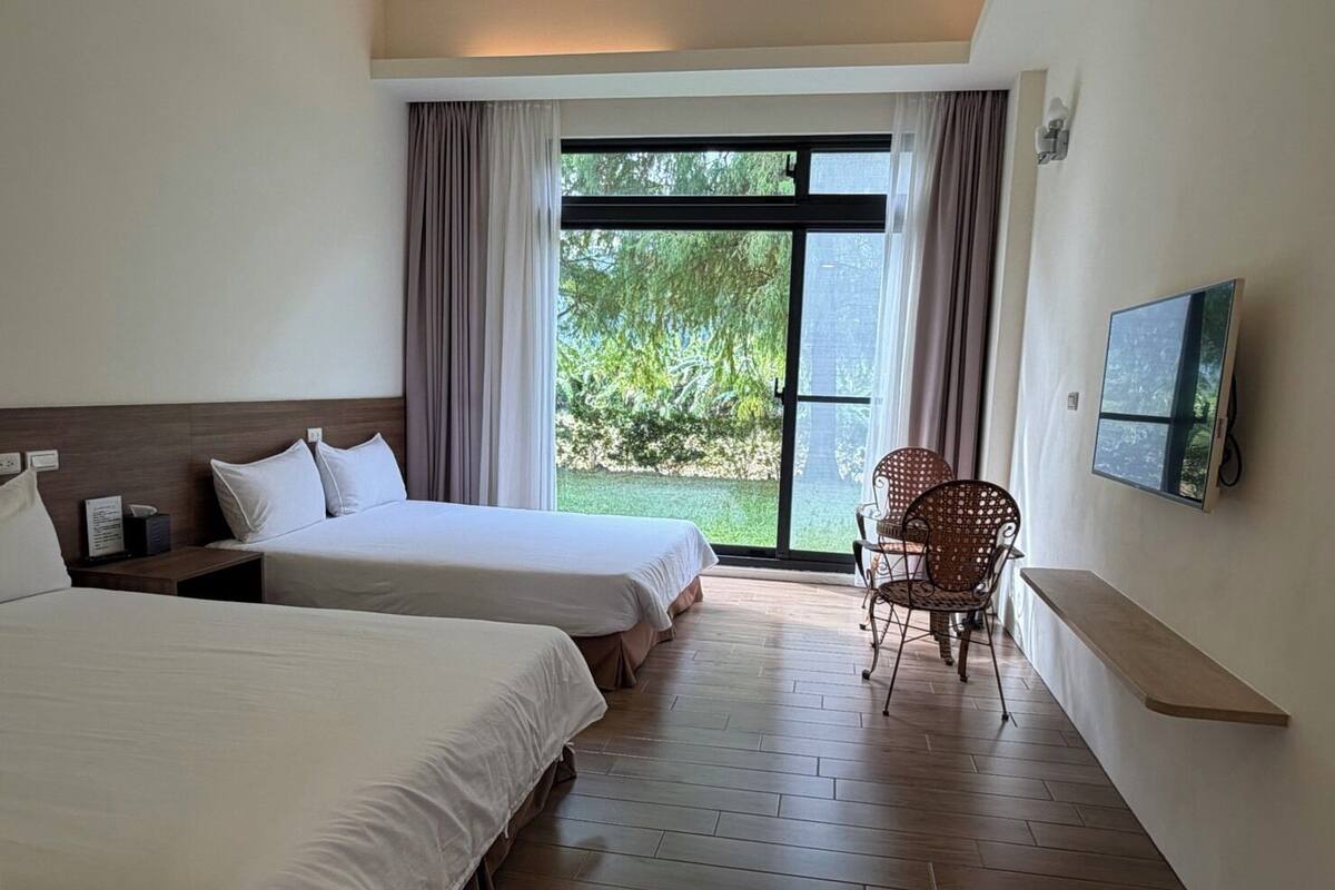 Habitación de hotel con dos camas, televisión, una silla y vista a zonas verdes a través del ventanal.