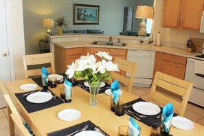 A dining area with a wooden table set for four, surrounded by chairs. A vase with white flowers is placed in the center.