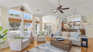 A spacious living room with a large window, a ceiling fan, and a dining area with a view. - Sound & Sol: Soundfront, Private Pool, Hot Tub (Corolla)
