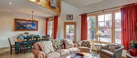 A spacious living room with a high ceiling, a dining area, and large windows with red curtains.