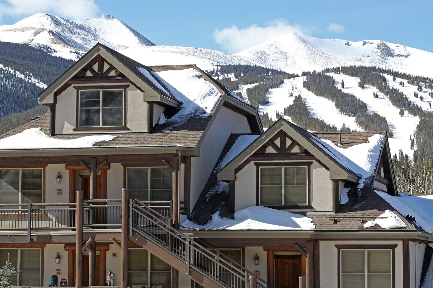 A two-story lodge with a balcony and a staircase leading to the upper floor, set against a backdrop of snow-covered mountains.