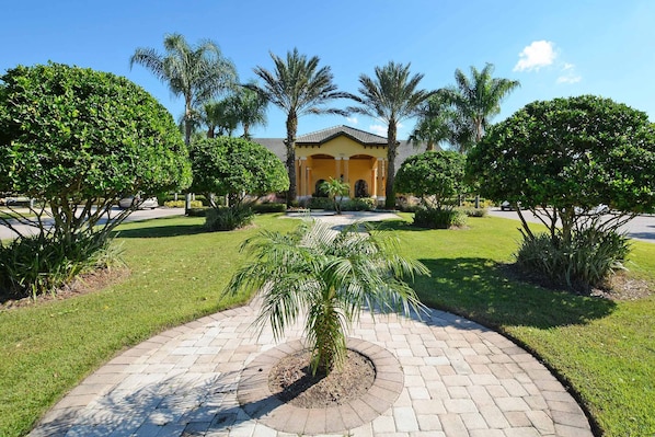 A courtyard with a paved walkway, palm trees, and a building with a yellow facade and arched entrance.