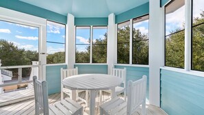 Un espace extérieur abrité, avec une table et des chaises blanches, de grandes fenêtres et une vue sur des arbres et un bâtiment.