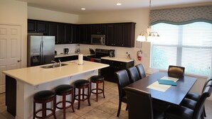 A modern kitchen with dark cabinets, a central island with stools, a dining table with chairs, and a large window with blinds. - Luxurious Home with Private Pool Upstairs  Games Room. 928 (Davenport)