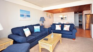 A living room with two blue sofas, a wicker coffee table, and a ceiling fan. - Cozy 1 Bedroom Oceanfront Condo with Free WiFi, Beach Decor, an Outdoor Pool, and a Breathtaking View Located Midtown and Steps from the Beach! (Ocean City)