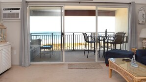 A room with a sliding glass door leading to a balcony with a view of the beach. There is a small table with chairs and a sofa. - Cozy 1 Bedroom Oceanfront Condo with Free WiFi, Beach Decor, an Outdoor Pool, and a Breathtaking View Located Midtown and Steps from the Beach! (Ocean City)