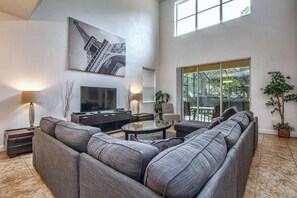 A modern living room with a large sectional sofa, a flat-screen TV, a glass coffee table, and a large window offering a view of the outdoors.