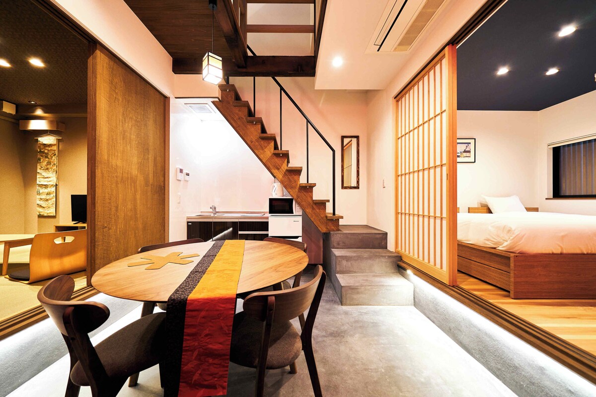 A modern hotel room with a wooden staircase, a dining area with a round table, and a bed with a large window.