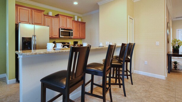 A modern kitchen with a central island, wooden cabinets, a stainless steel refrigerator, and a microwave. There are three bar stools and a potted plant on a small table.