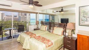 A bedroom with a large bed, a nightstand, a dresser, and a balcony with a view of the city and ocean.