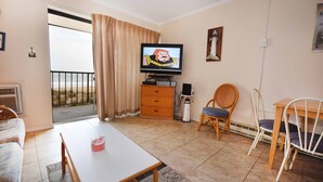 Una sala de estar con televisor, una mesa, sillas, un mueble bajo y vista a la playa a través de una puerta corrediza.