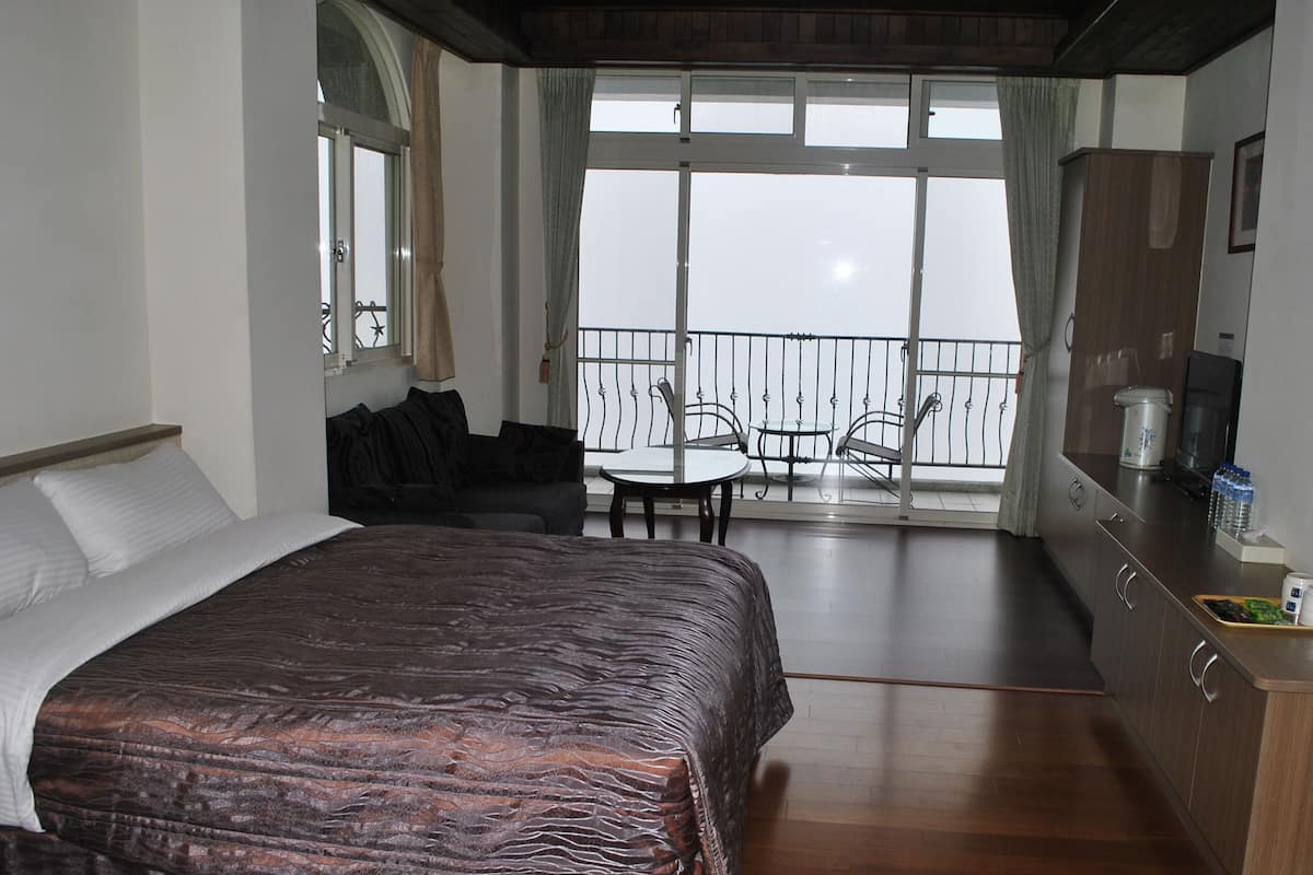 A hotel room with a bed, a desk, a chair, a balcony with a table and chairs, and a fan.