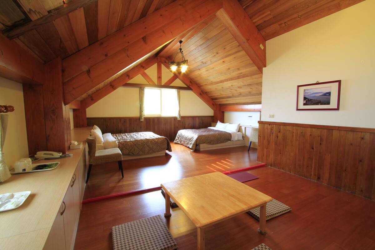 A cozy attic bedroom with wooden beams, two beds, a wooden table, and a view of the outdoors.