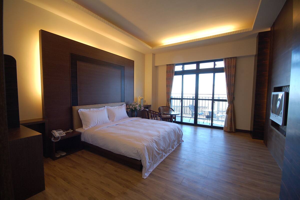 A hotel room with a large bed, wooden flooring, a nightstand with a phone, a window with curtains, and a balcony with chairs.