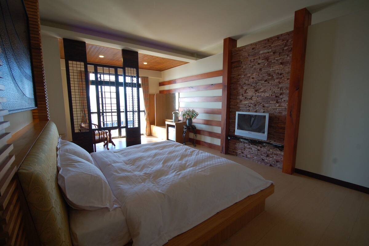 A bedroom with a large bed, a TV mounted on a brick wall, and a desk with a chair.