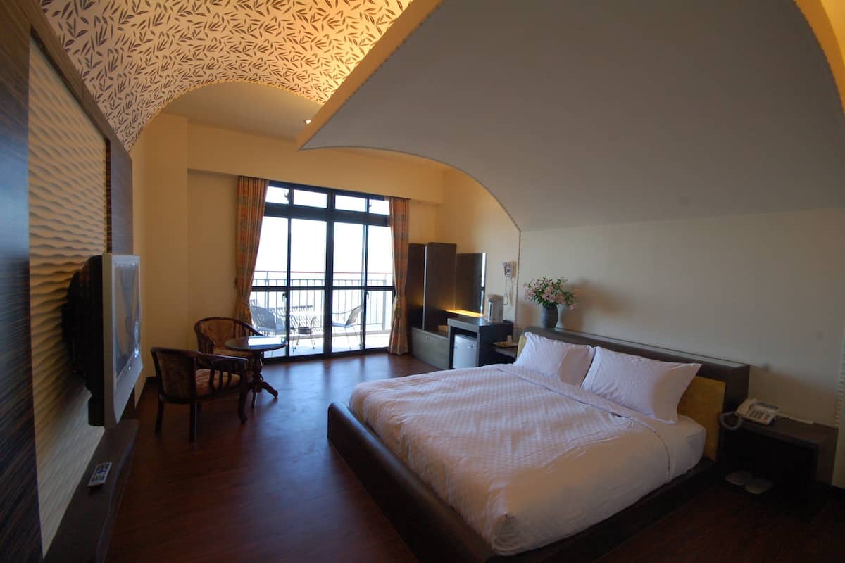A hotel room with a large bed, a desk, a chair, a TV, and a balcony with a view.