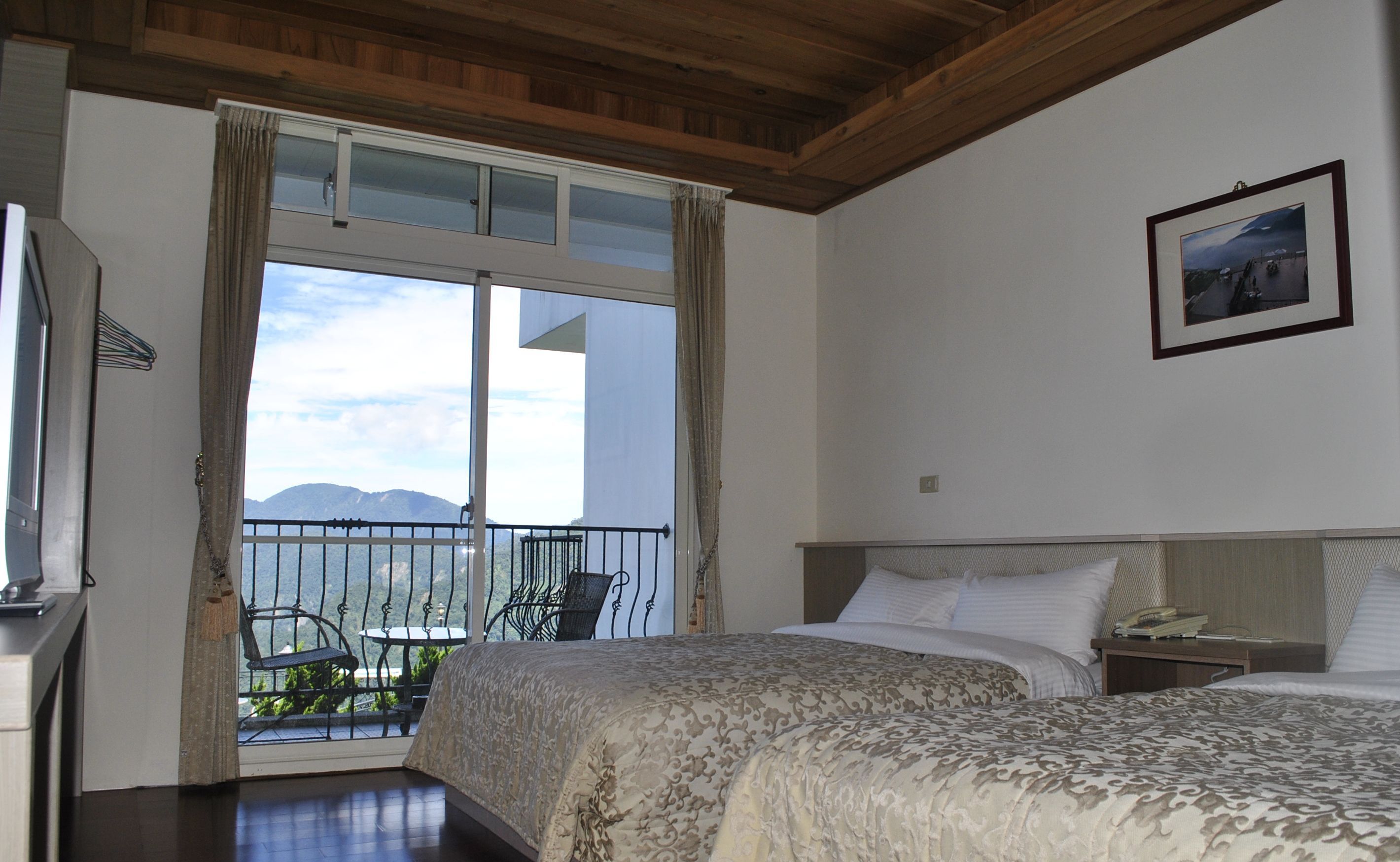 A hotel room with a large bed, a bedside table, a chair, and a balcony with a view of the mountains.