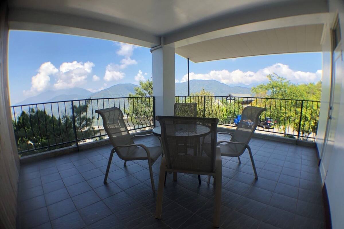 A balcony with a table and chairs, offering a view of mountains and trees.
