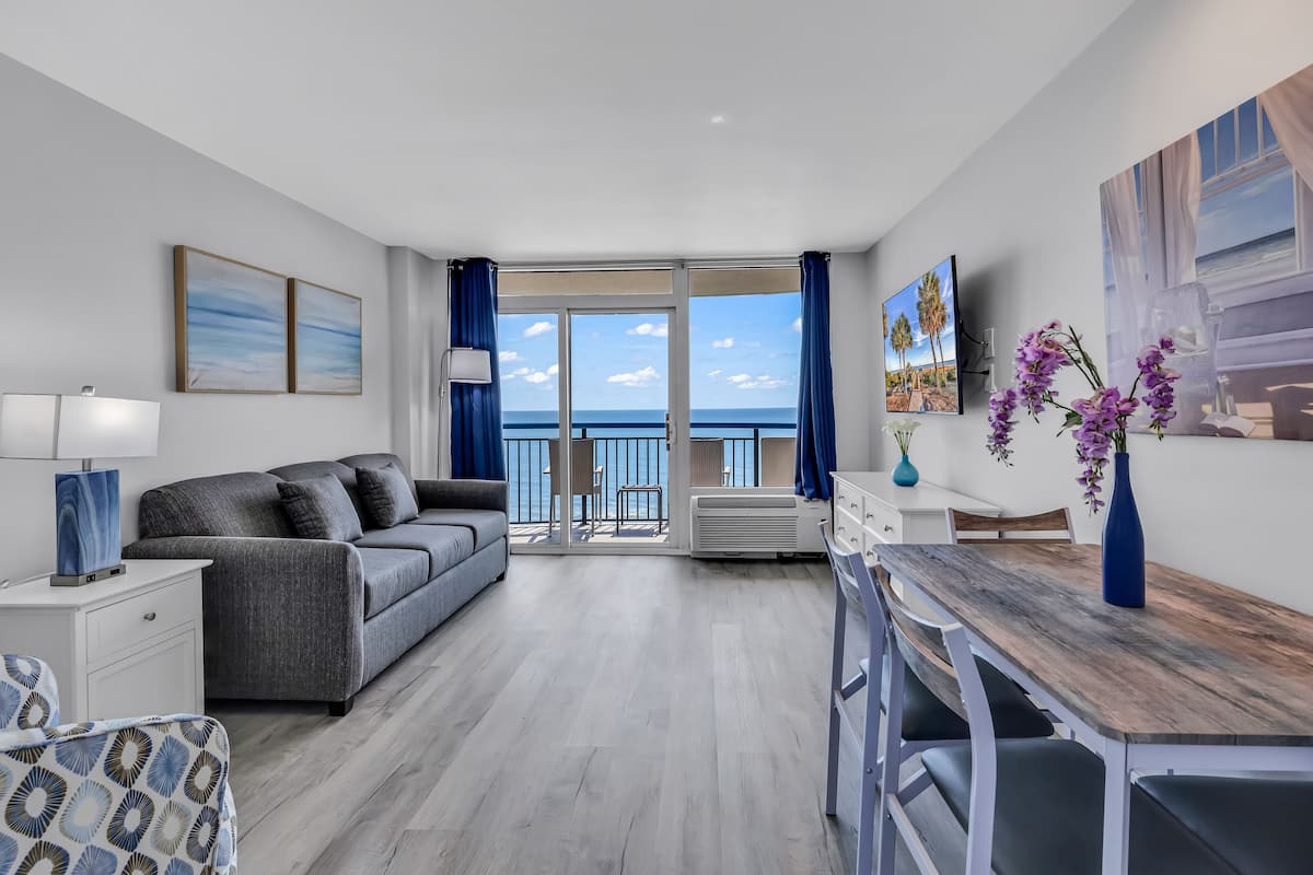 A modern living room with a grey sofa, a wooden dining table, and a balcony with a view of the sea.