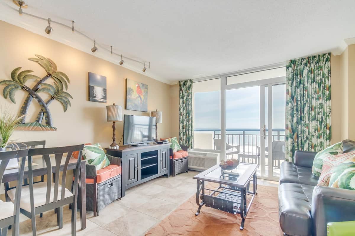 A living room with a television, dining table, and chairs, a sofa, and a view of the ocean through large windows.