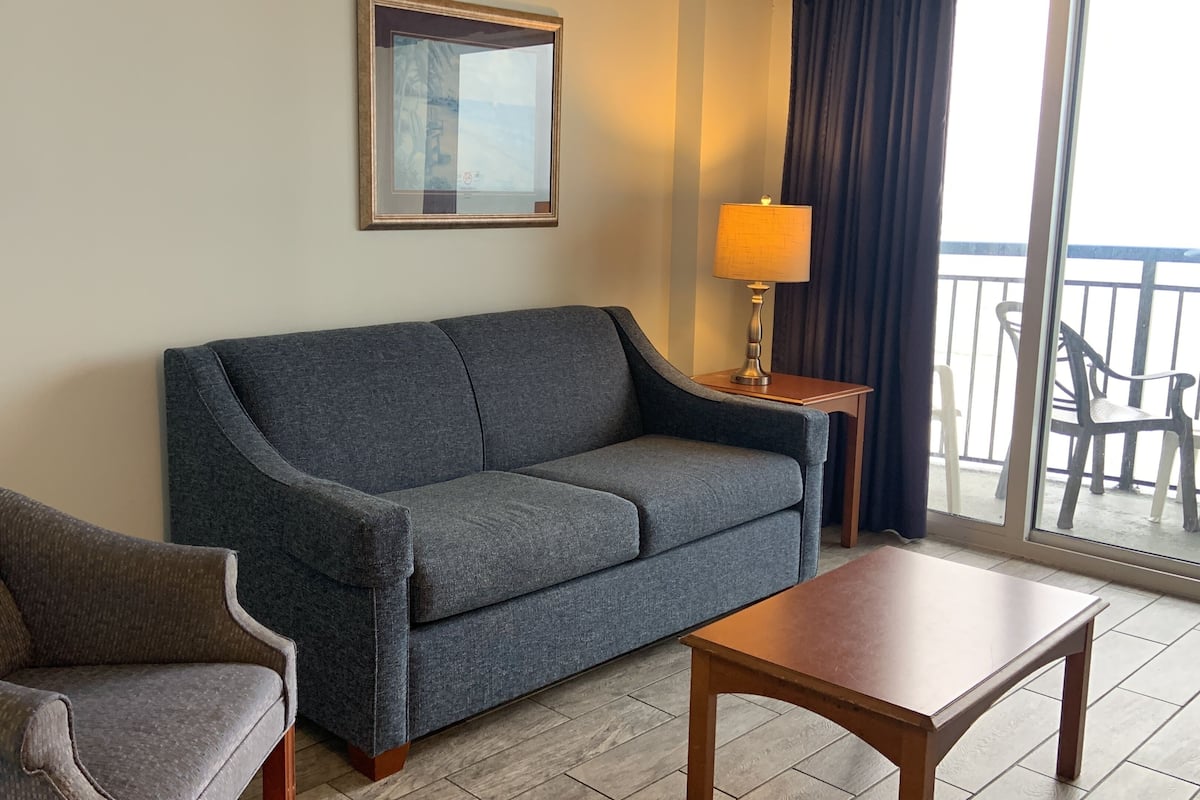 A hotel room with a grey sofa, a brown armchair, a wooden coffee table, a lamp, and a view of the balcony.