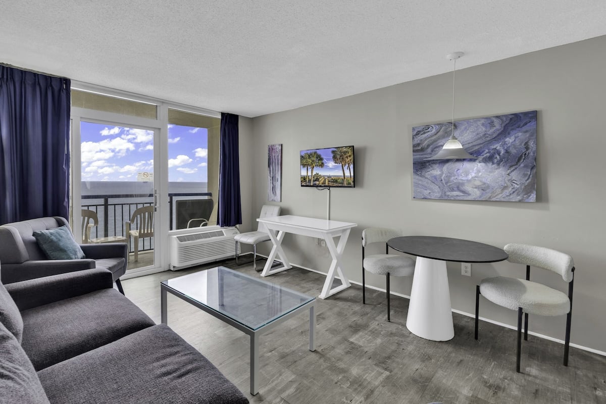 A modern living room with a glass coffee table, grey sofas, a white side table, and a dining area with a white table and chairs.
