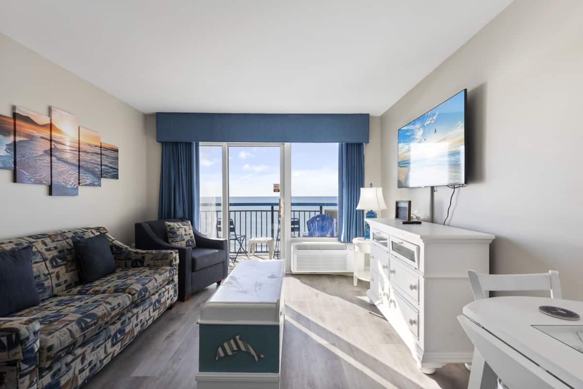 A modern hotel room with a balcony, a sofa, a coffee table, a TV, and a dresser.