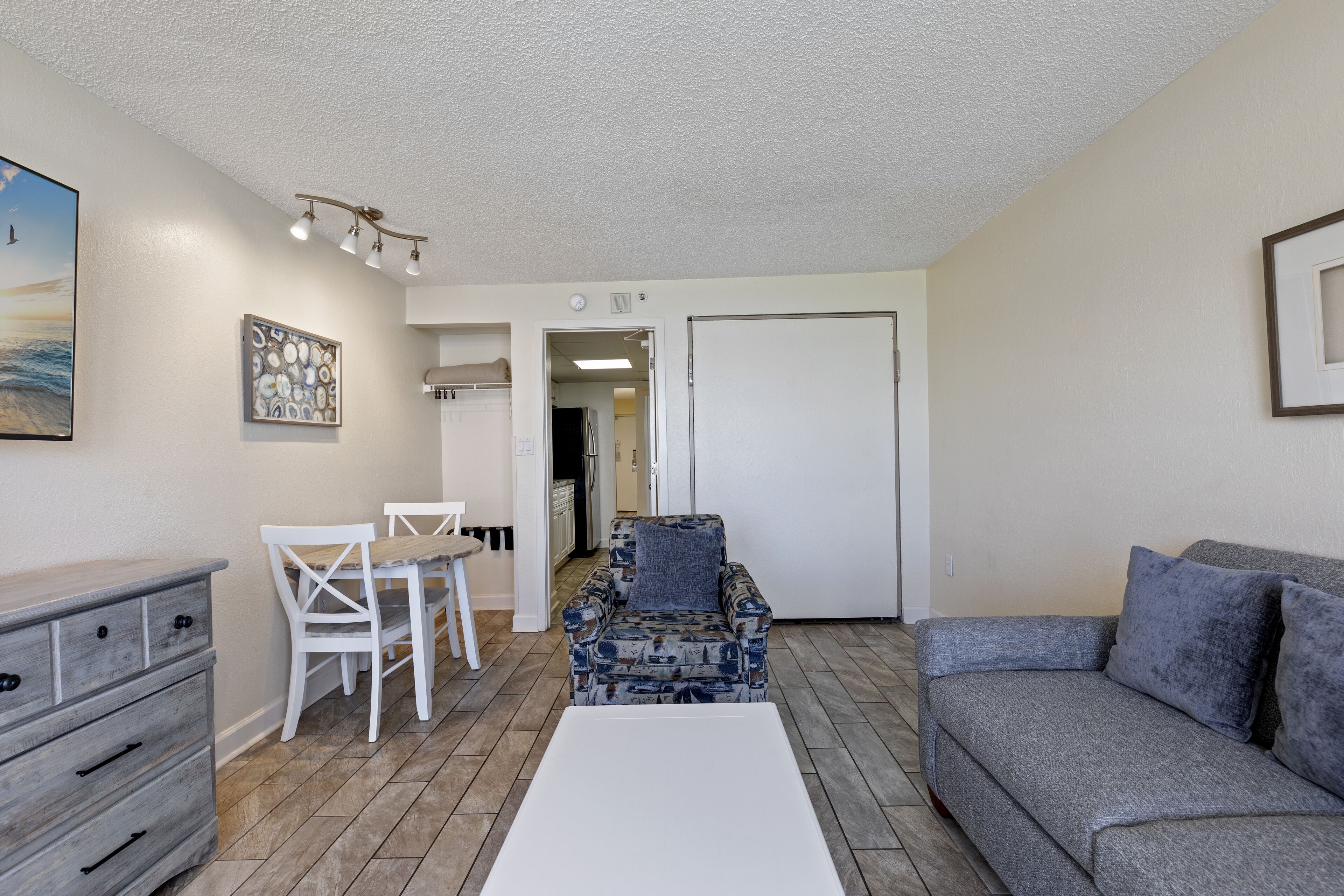 A living room with a gray sofa, a patterned armchair, a white coffee table, and a dining area with a white table and chairs.