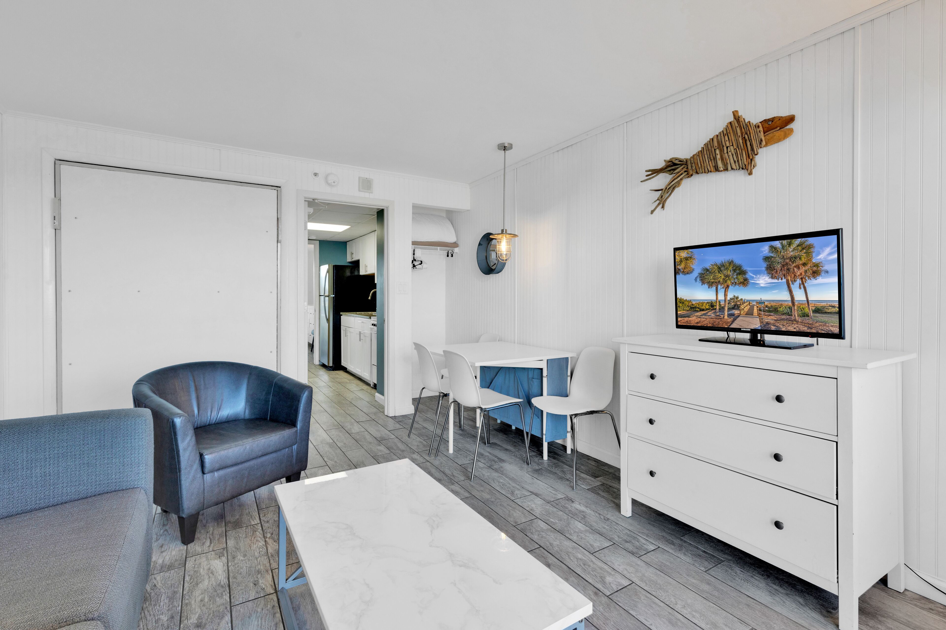 A modern living room with a white sofa, a blue armchair, a marble coffee table, a white TV stand with a flat-screen TV, and a dining area with a white table and chairs.