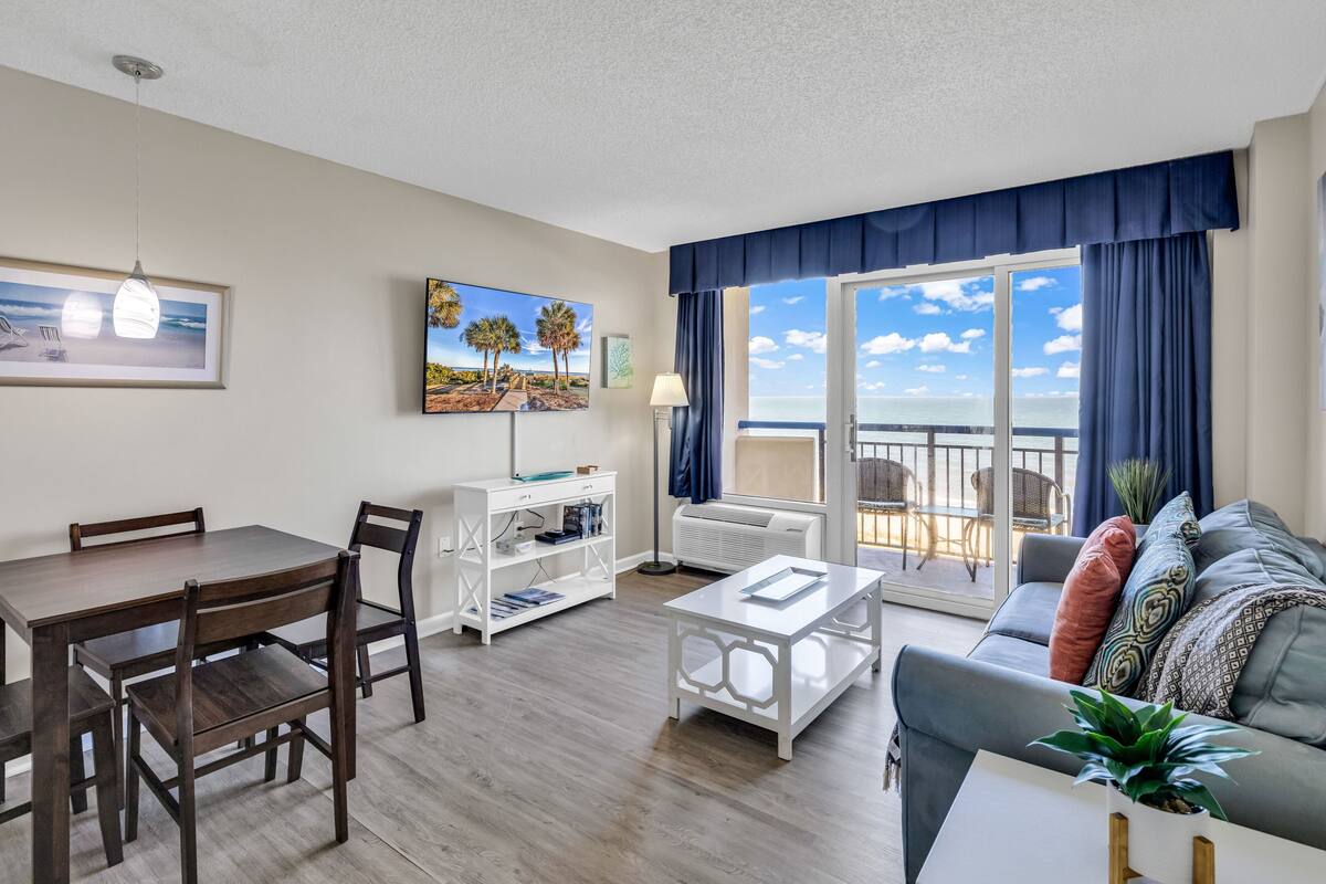 A modern living room with a dining area, a sofa, a coffee table, and a balcony with a view of the sea.