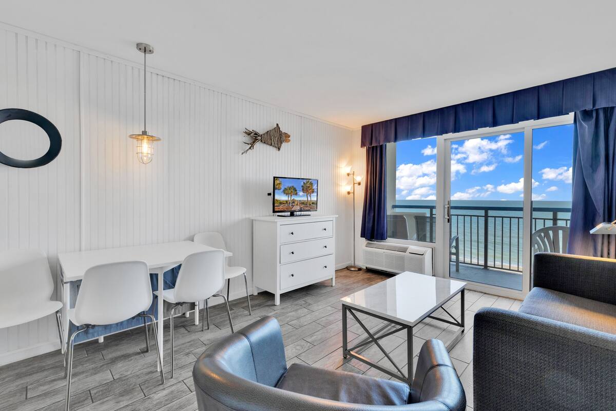 A modern living room with a dining area, a large window with a view of the sea, and a balcony.