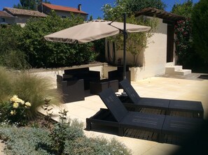 Eine Terrasse mit Gartenmöbeln, darunter eine Lounge-Liege und ein Tisch, unter einem großen Sonnenschirm.