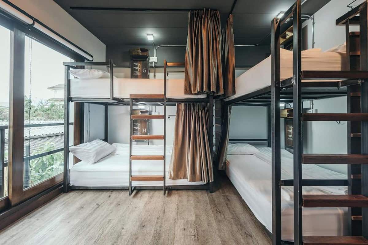 A bunk bed room with a wooden floor, a window with curtains, and a small shelf with books.