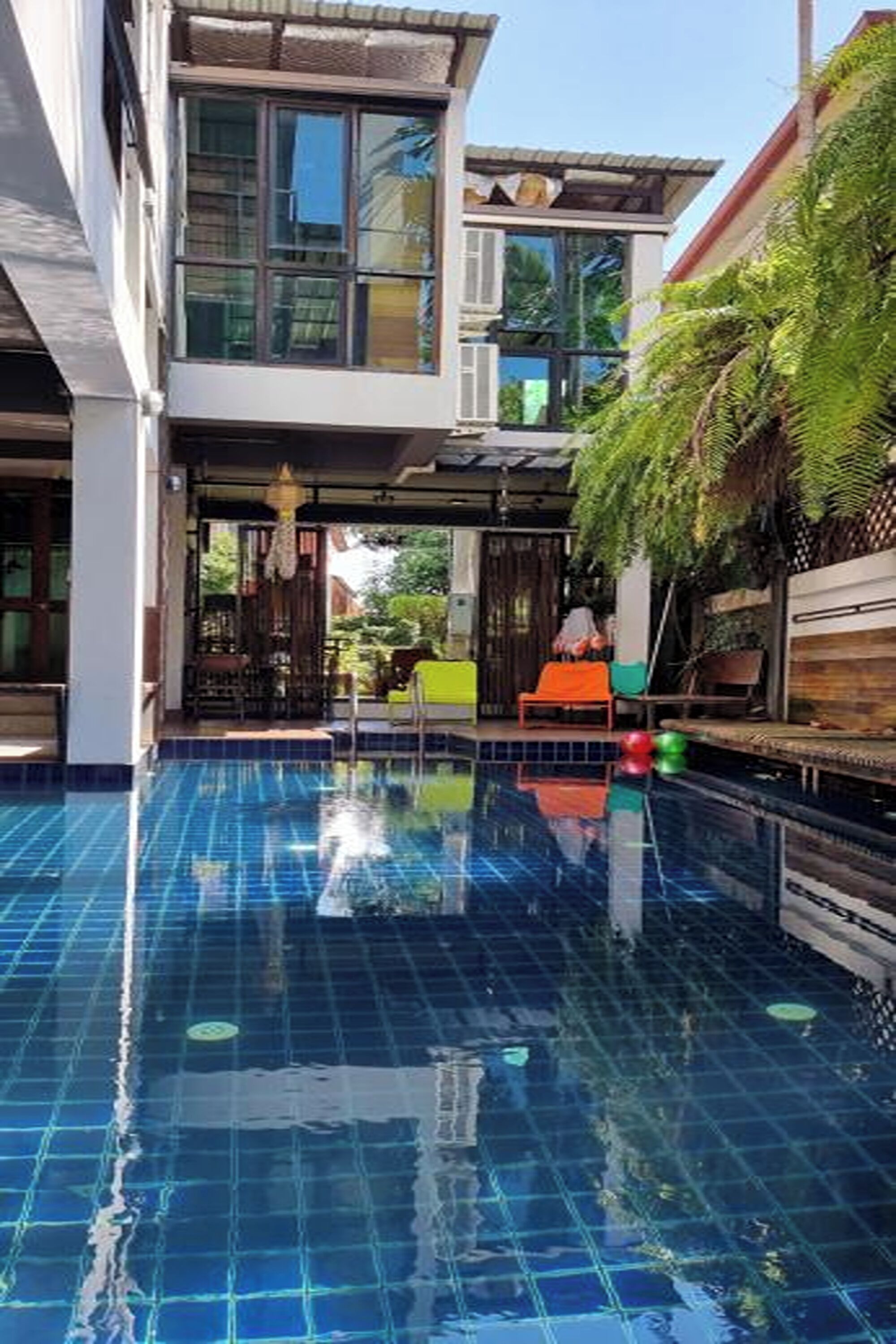 A swimming pool with tiled flooring, surrounded by a building with large windows and a covered area with seating.