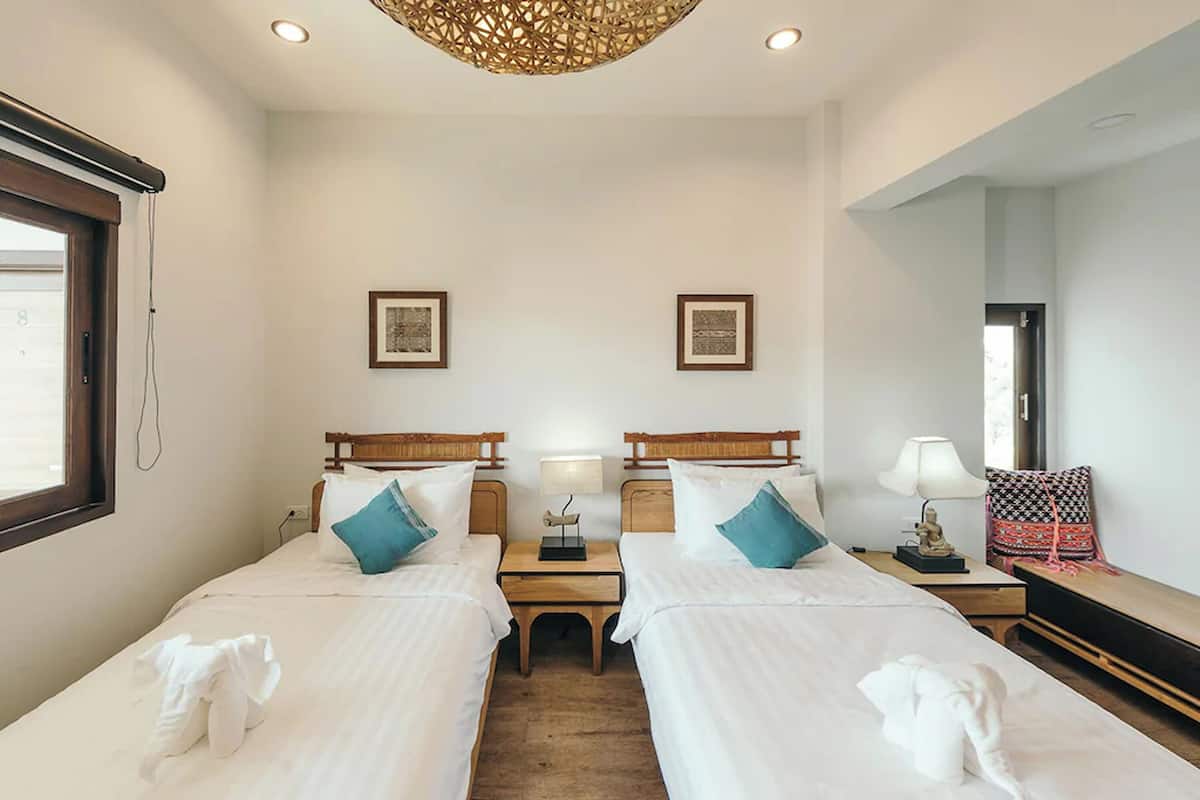 A hotel room with two single beds, wooden headboards, white bedding, teal pillows, a small bedside table with a lamp, and a large mirror on the right wall.