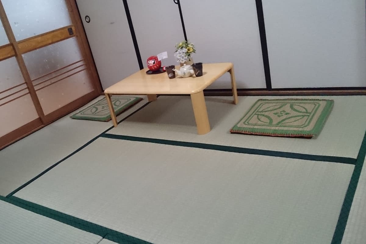 A tatami mat room with a low wooden table, a small red and white ceramic figure, a vase with flowers, and a green mat with a geometric pattern.