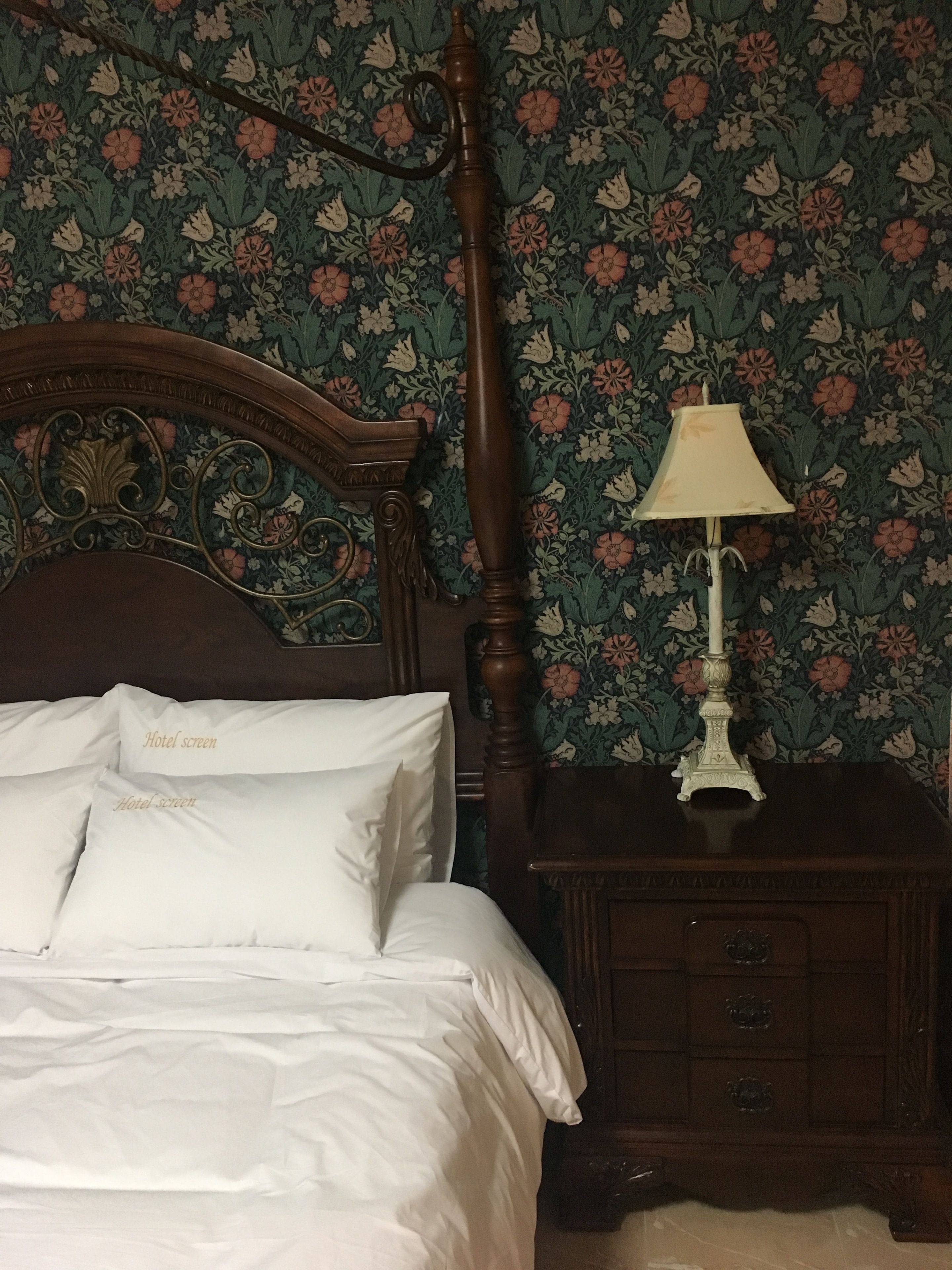 Una cama bien hecha con sábanas y almohadas blancas, con la etiqueta "Hotel screen". La habitación tiene papel tapiz floral y una mesita de noche de madera con una lámpara.
