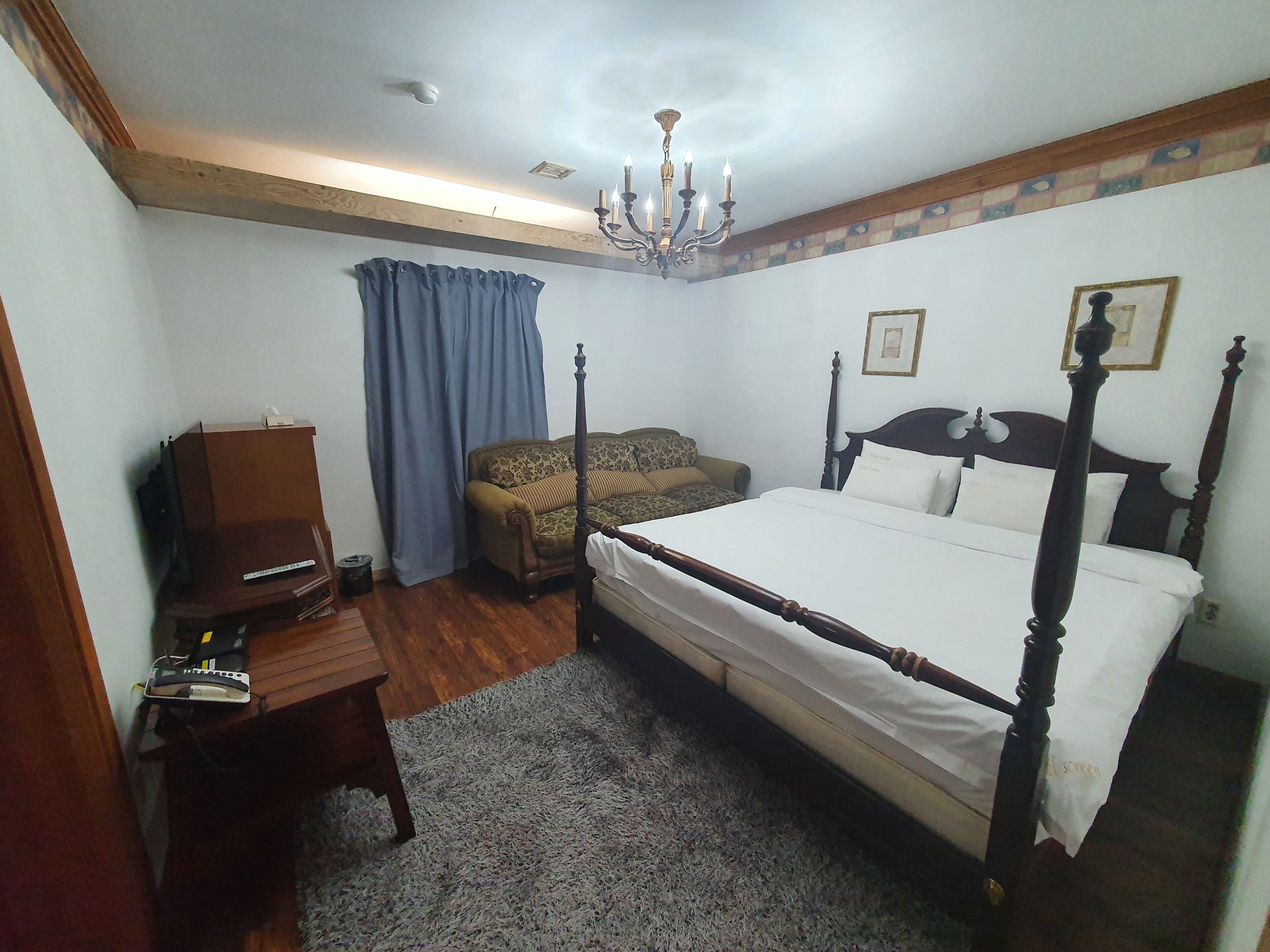 A bedroom with a four-poster bed, a wooden desk, a television, and a chandelier.