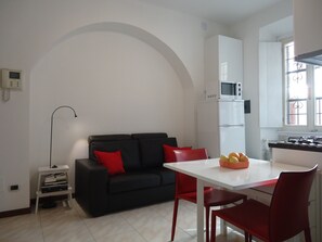Un ambiente compatto con un divano nero, sedie rosse, un tavolo bianco e un angolo cottura dotato di forno a microonde e piano cottura.