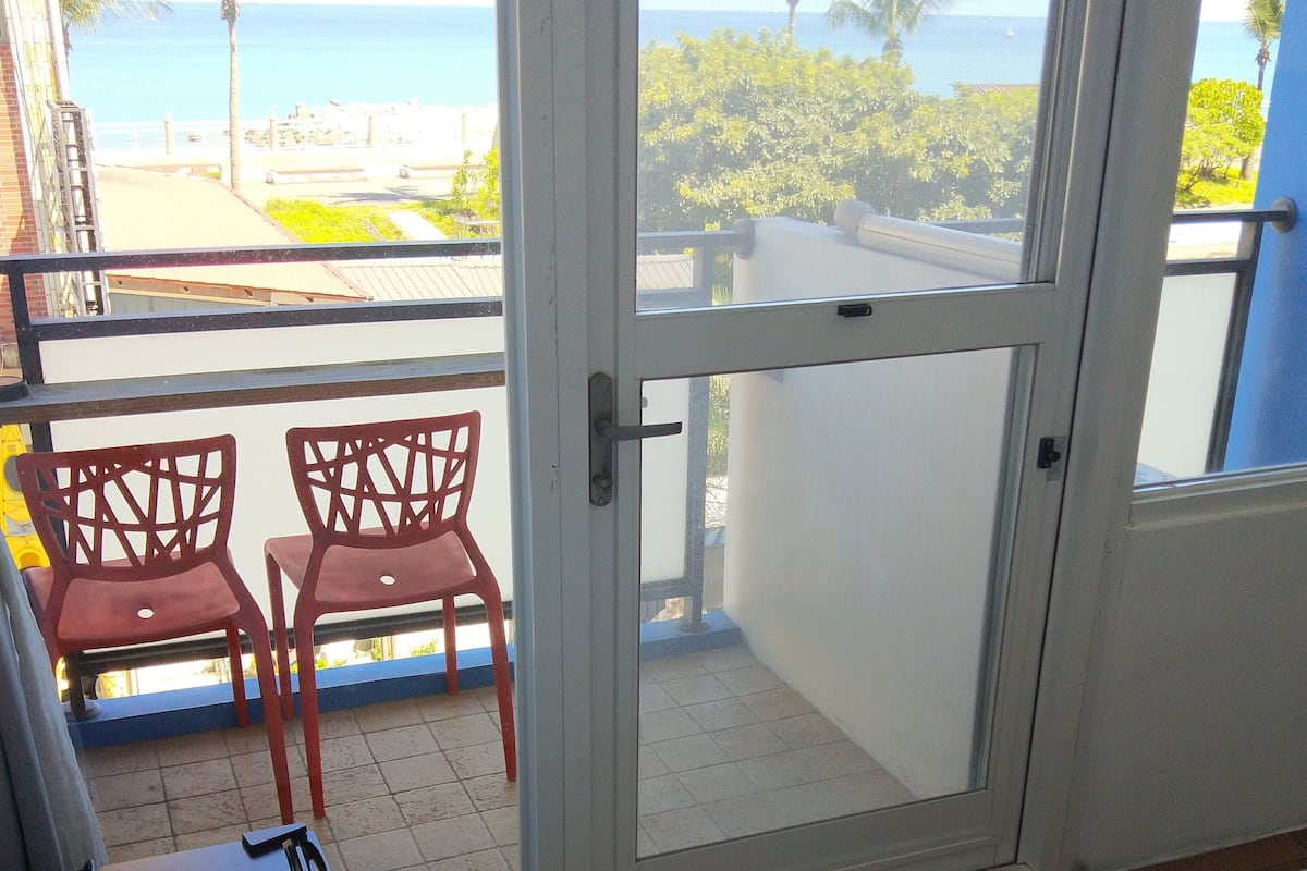 A room with a balcony overlooking a beach and ocean. There are two red chairs and a small table on the balcony.